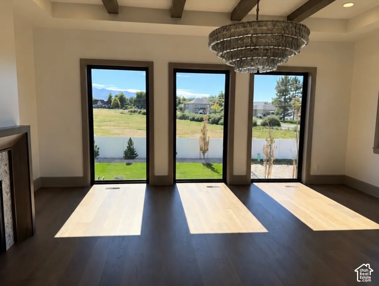 Hardwood flooring, three full-height windows, decorative overhead lighting fixture, exposed ceiling beams, and a fireplace with stone surround