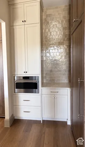 Built-in stainless steel microwave, white shaker-style cabinetry, wood-finish flooring, and a tiled accent wall