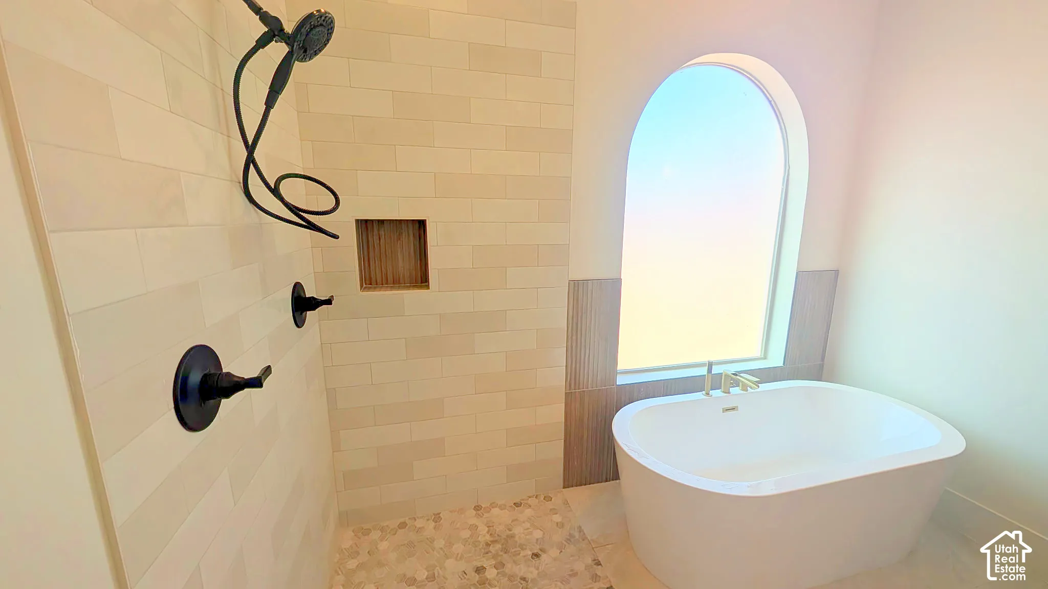 Freestanding soaking tub, arched window, matte black wall-mounted shower fixtures, tiled shower niche, and custom stone tile shower flooring