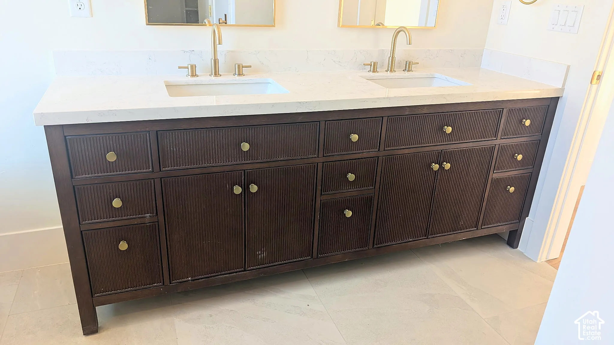 Double vanity featuring a textured dark wood cabinet, white stone countertop with dual integrated sinks, and brass hardware