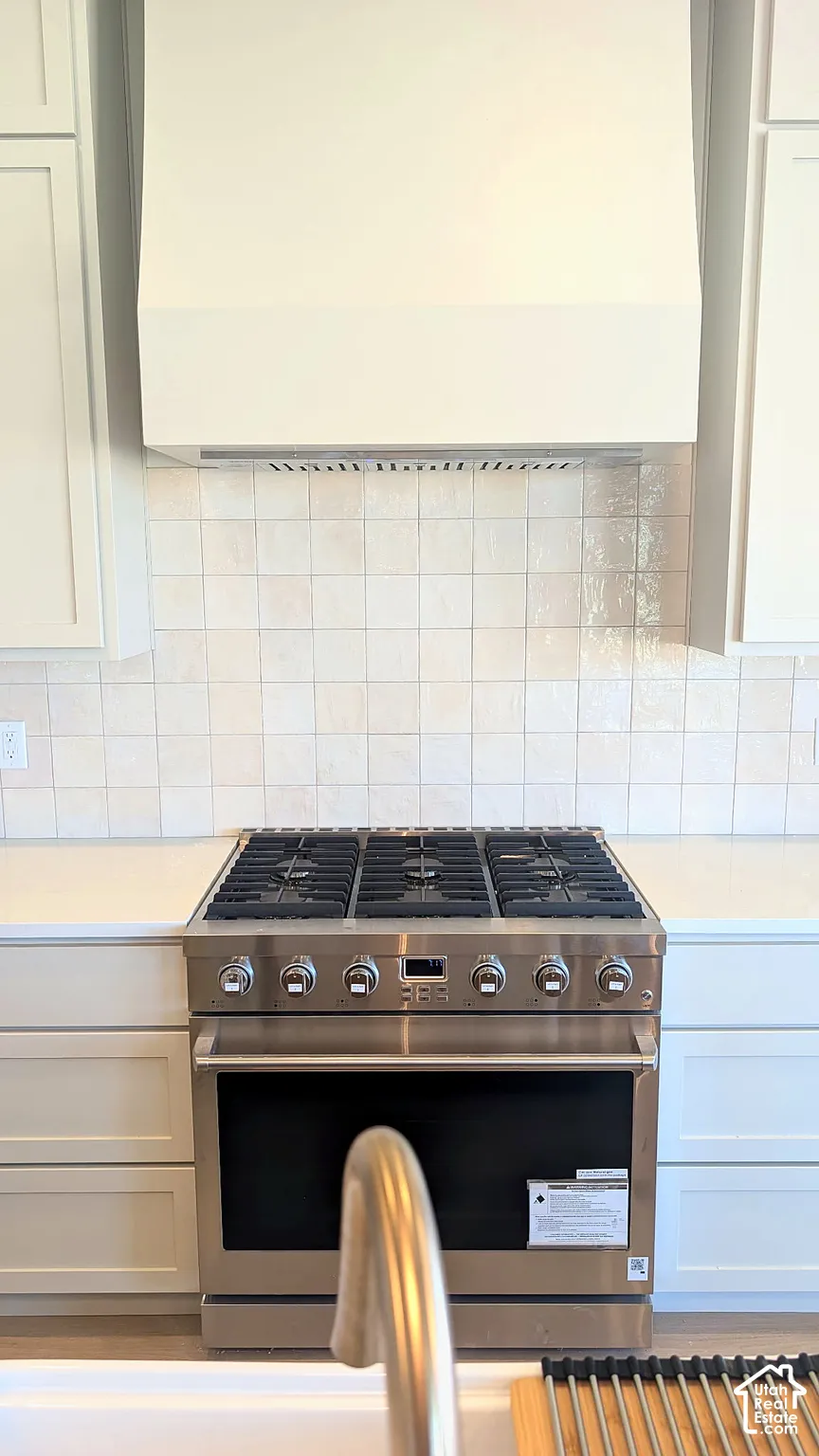 Stainless steel six-burner gas range, custom-built vent hood, square-tile backsplash, and light-toned solid surface countertops featuring Shaker-style cabinetry