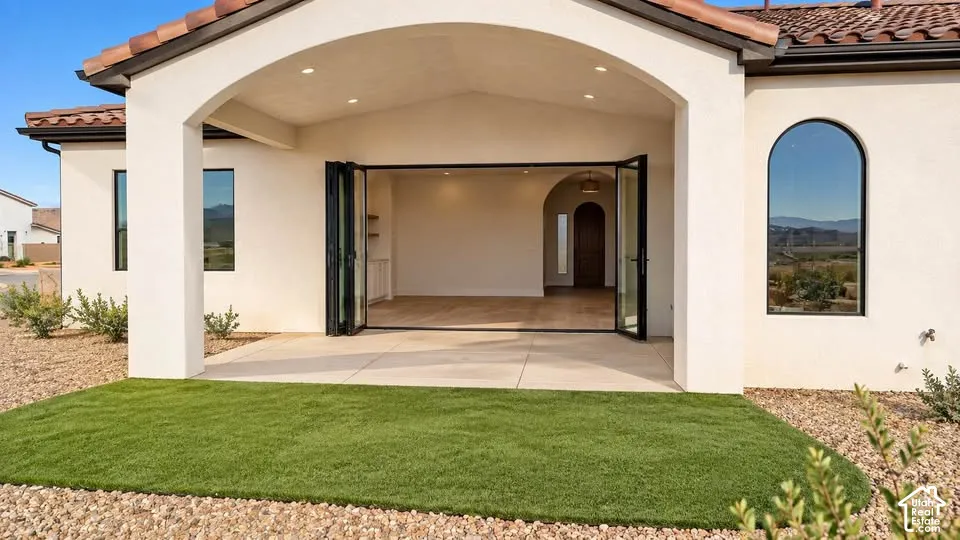 Covered patio featuring an arched stucco exterior, multi-panel folding glass doors, concrete flooring, recessed lighting, and synthetic turf lawn