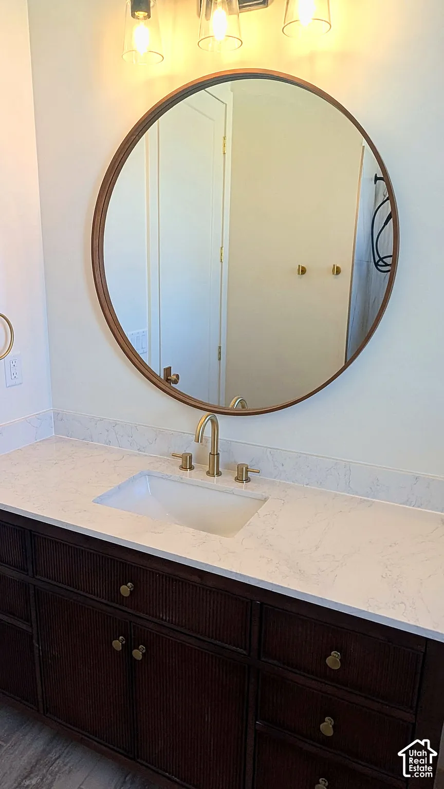 Dark wood-toned vanity featuring a textured facade, stone-finish countertop with an integrated rectangular sink, brass-toned widespread faucet, and circular framed mirror
