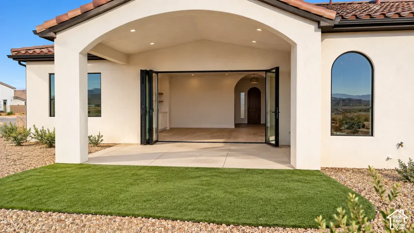 Covered patio with recessed lighting, bi-fold glass doors, arched entryways, and a manicured artificial turf lawn