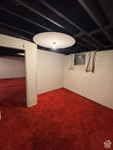 Red carpet flooring, white painted walls, exposed black ceiling joists, flush-mount ceiling light fixture, and a window with a sheer curtain