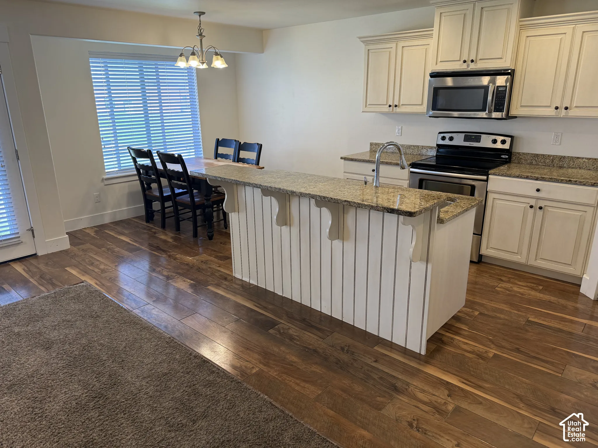 Wood-finish flooring, L-shaped kitchen with granite countertops, stainless steel appliances, kitchen island with beadboard paneling, and a dining area with a chandelier