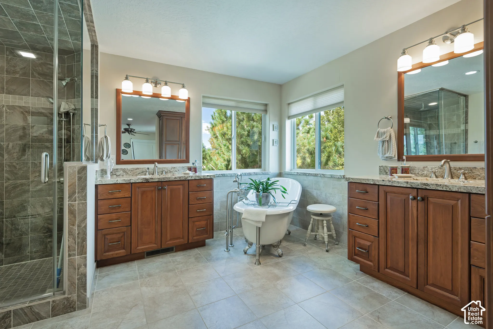 Dual wood-finish vanities with stone countertops, a freestanding clawfoot tub, a glass-enclosed shower with tiled walls, and large windows