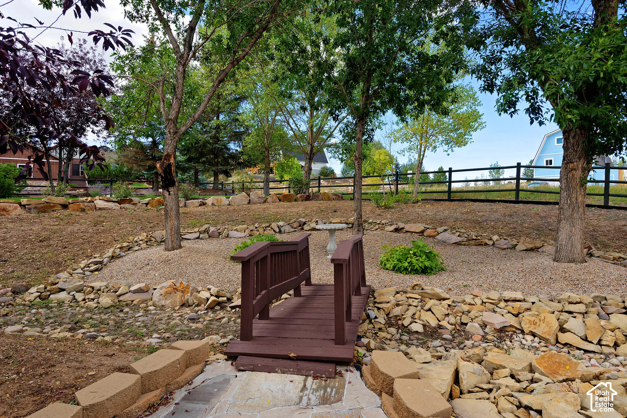 Landscaped yard featuring a decorative bridge, mature trees, natural stone accents, and a wood fence