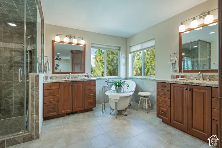 Dual wood-finish vanities with stone countertops, a freestanding clawfoot tub, a glass-enclosed shower with tiled walls, and large windows