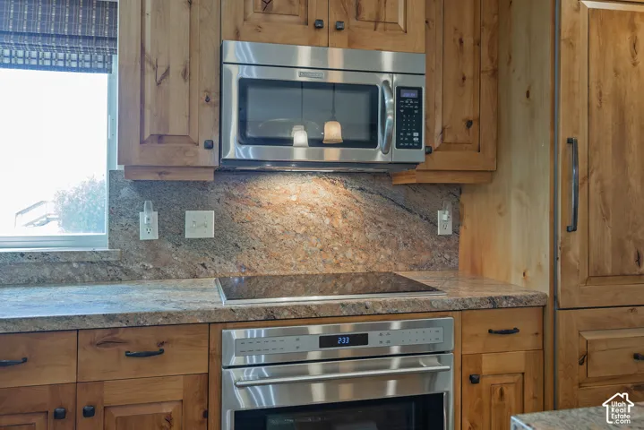 Wood cabinetry, stainless steel microwave, cooktop, built-in oven, and stone-finish countertops
