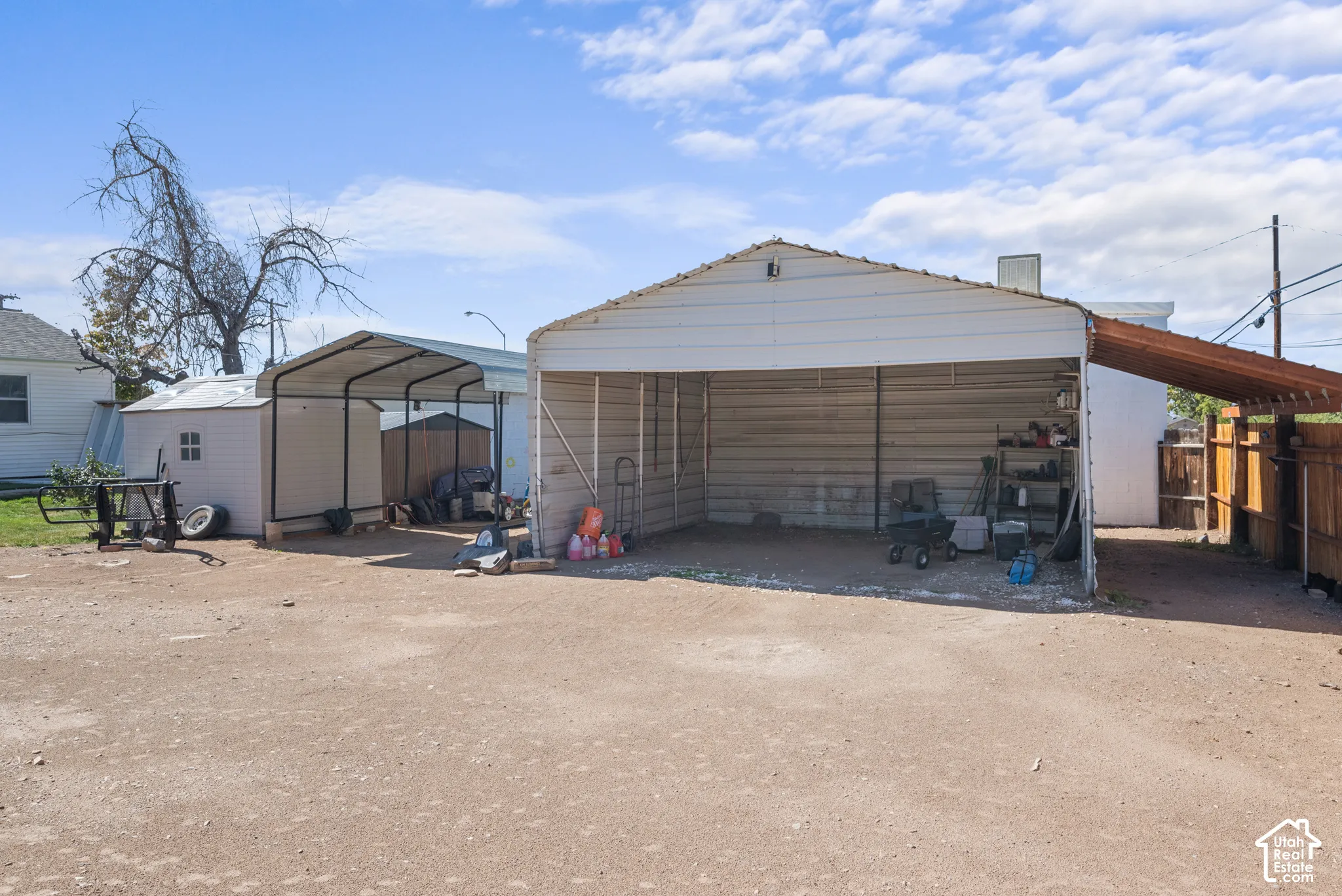 Metal carport structure, gravel ground cover, a second covered structure, and a wood-slat fence