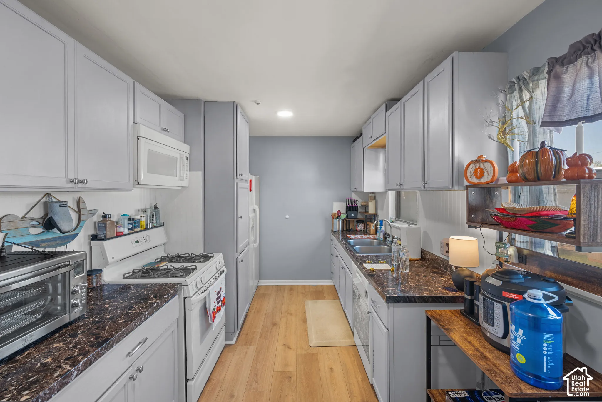 Galley kitchen featuring light gray cabinetry, dark wood-finish countertops, white appliances, wood-finish flooring, and a stainless steel double basin sink