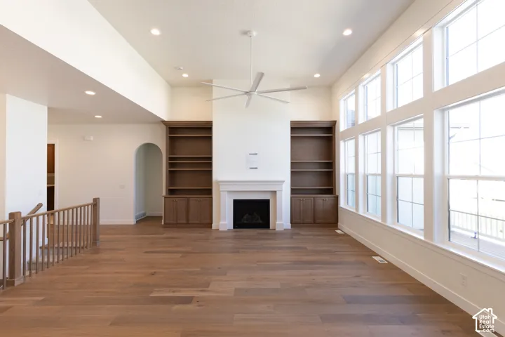 Expansive living area with wood-finish flooring, a white mantel fireplace, built-in shelving, vaulted ceilings, and large multi-pane windows