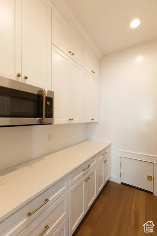 White shaker-style cabinetry with gold-tone hardware, stainless steel built-in microwave, light-toned countertop, and wood-finish flooring