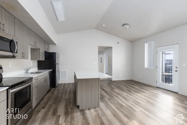 Vaulted ceiling above wood-finish flooring, featuring a kitchen island with a white countertop, stainless steel appliances, and light-toned cabinetry