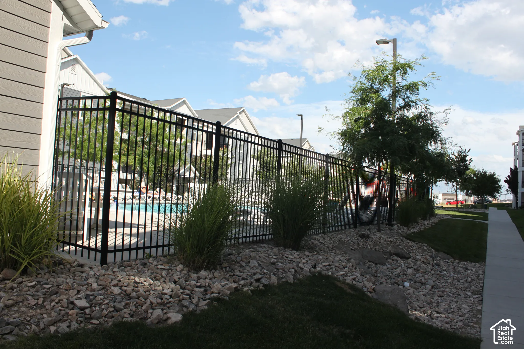 Outdoor swimming pool with surrounding fencing, adjacent to a sidewalk and landscaped rock and grass areas