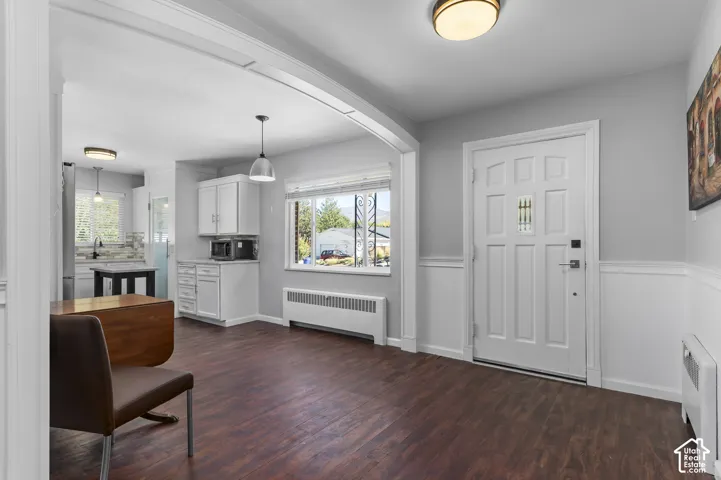 Wood-finish flooring throughout, white paneled entry door with decorative glass, and a kitchen featuring white cabinetry and pendant lighting