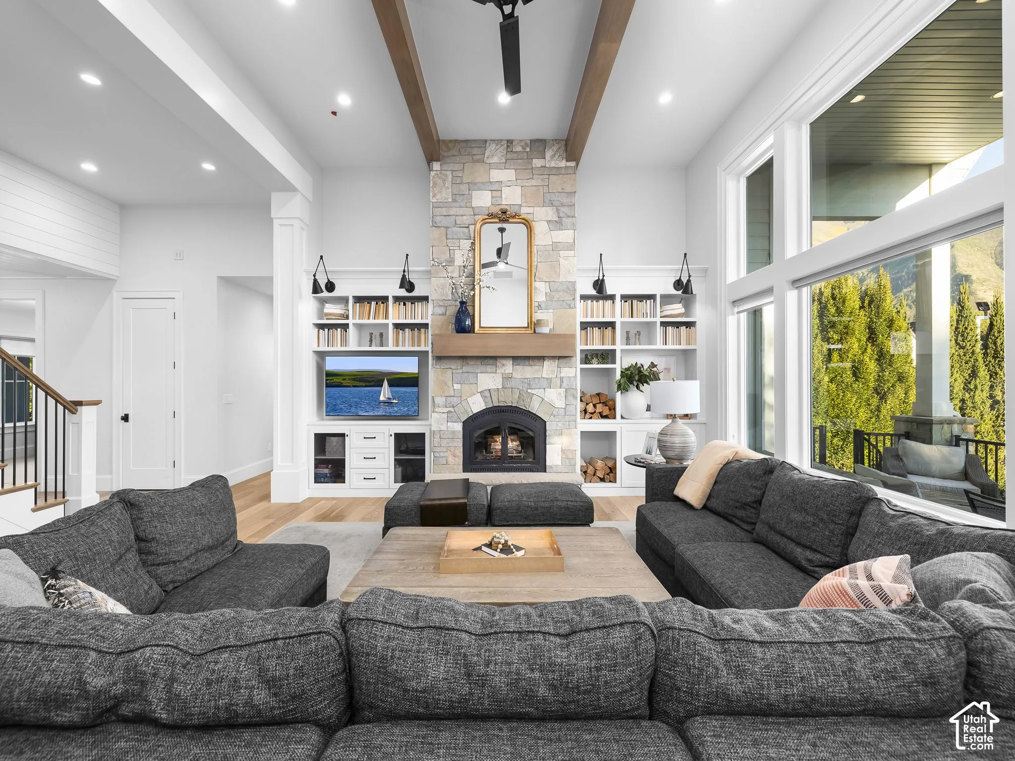 Vaulted ceilings with exposed wood beams, floor-to-ceiling stone fireplace, integrated built-in shelving, large windows, and light wood-finish flooring