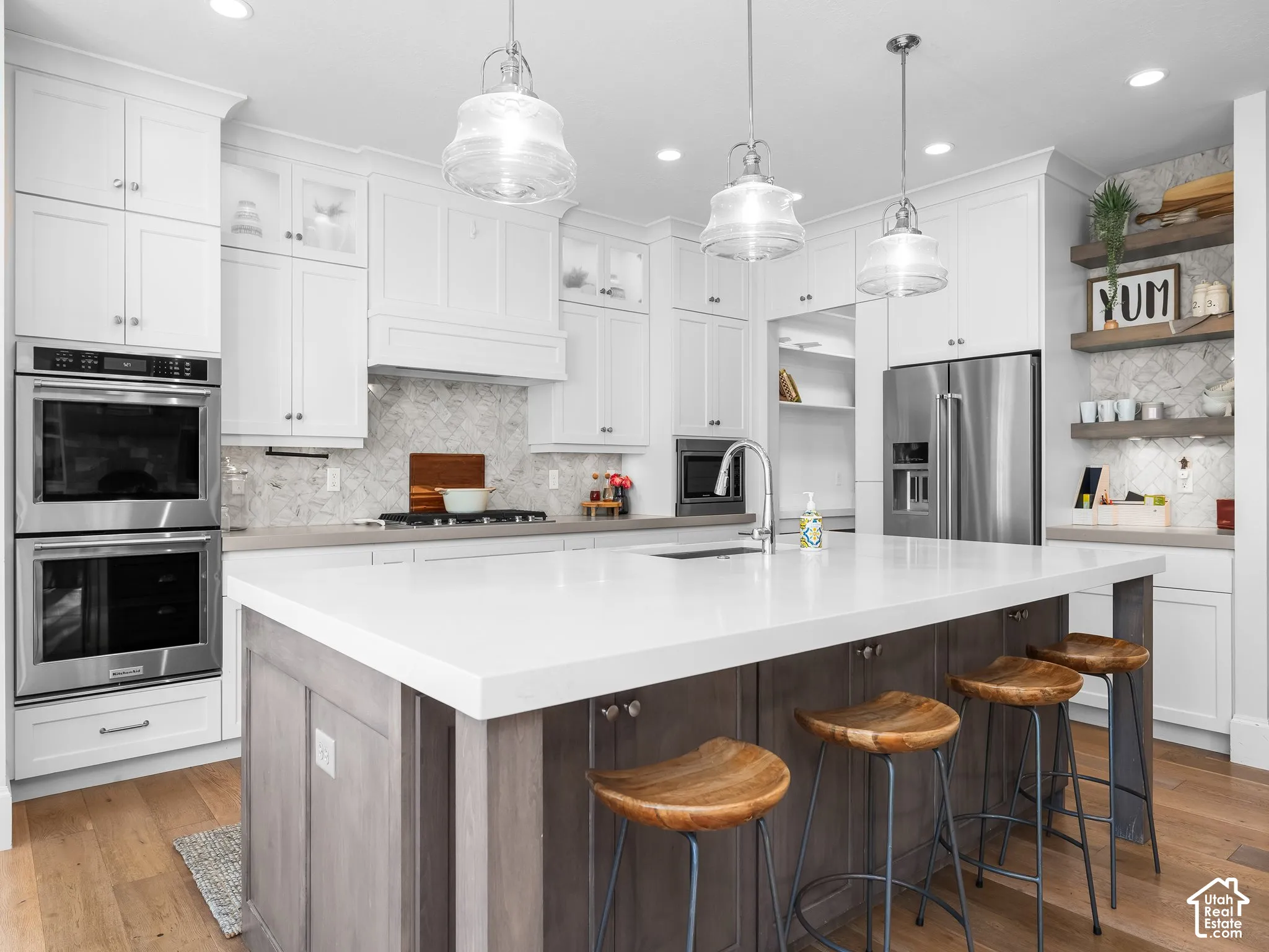 White shaker cabinetry with clear glass accents, stainless steel double wall ovens, a large kitchen island with a white countertop and seating, and wood-finish flooring