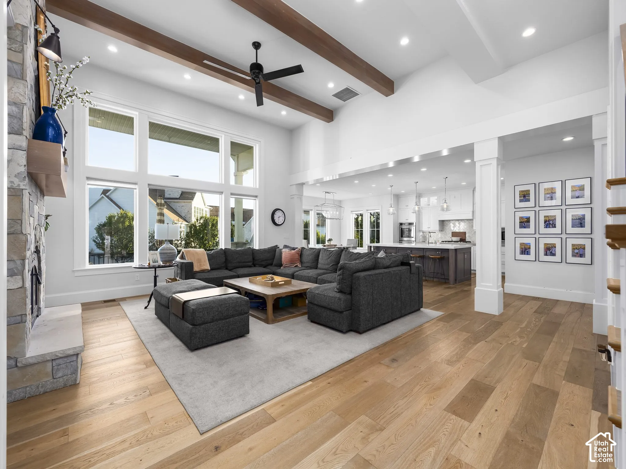High ceilings with exposed wood beams, stone-clad fireplace with wood mantel, light wood-finish flooring, recessed lighting, and expansive windows