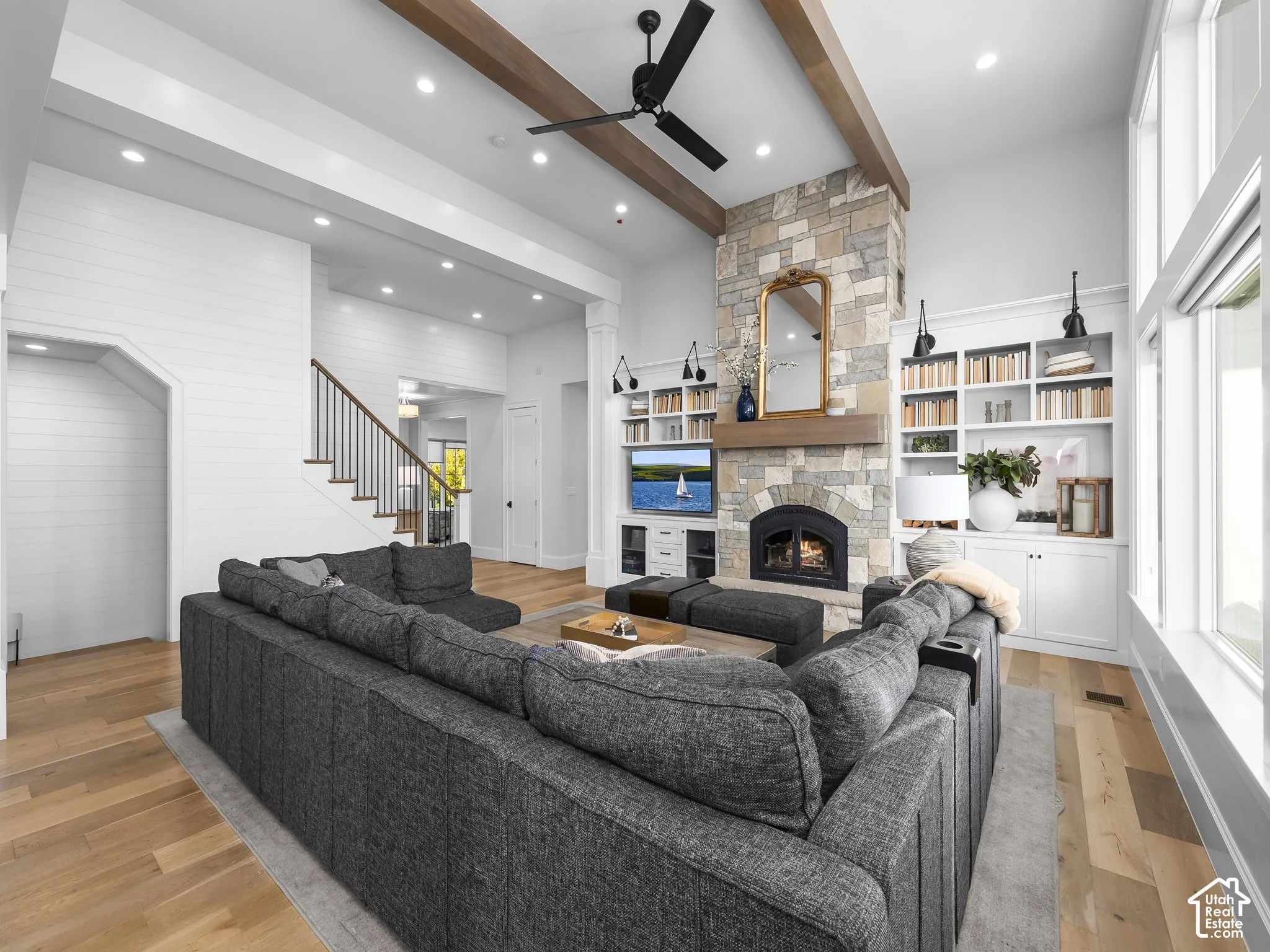 Spacious living area featuring a stone-clad fireplace, built-in shelving, wood-finish flooring, high ceilings with exposed beams, and a staircase with a wood handrail