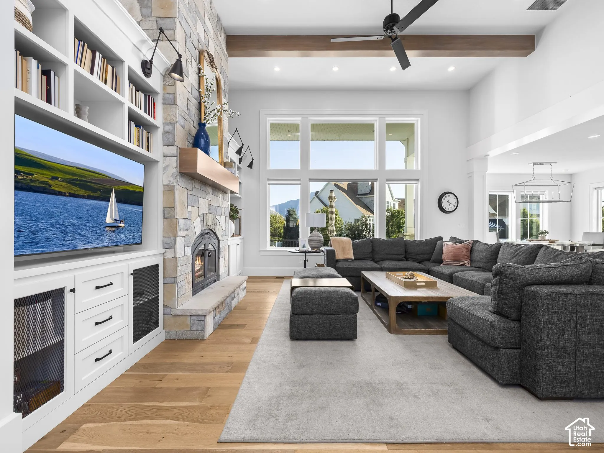 Vaulted ceiling with exposed beam, stone-faced fireplace, built-in shelving, large multi-pane windows, and wood-finish flooring