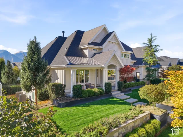 Gable roofline with dormer windows, stone and siding exterior, covered entry porch, manicured lawn, and multi-level landscaping