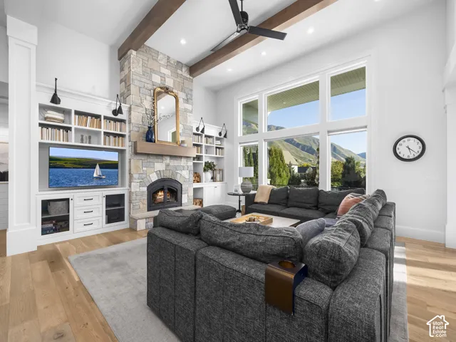 Vaulted ceiling with exposed wood beams, stone-clad fireplace with wood mantle, built-in cabinetry with integrated shelving, large multi-pane windows, and wide plank wood-finish flooring