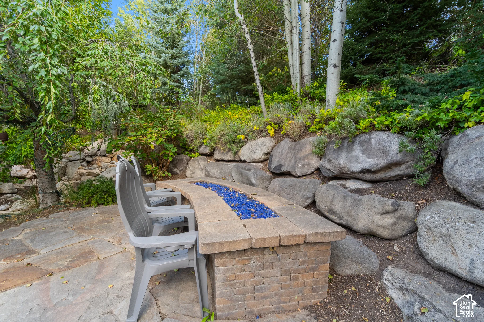 This stone patio has a built-in fire pit with blue glass, surrounded by natural rock retaining walls and mature trees, it sits next to the waterfall and overlooks the outdoor kitchen and seating areas.