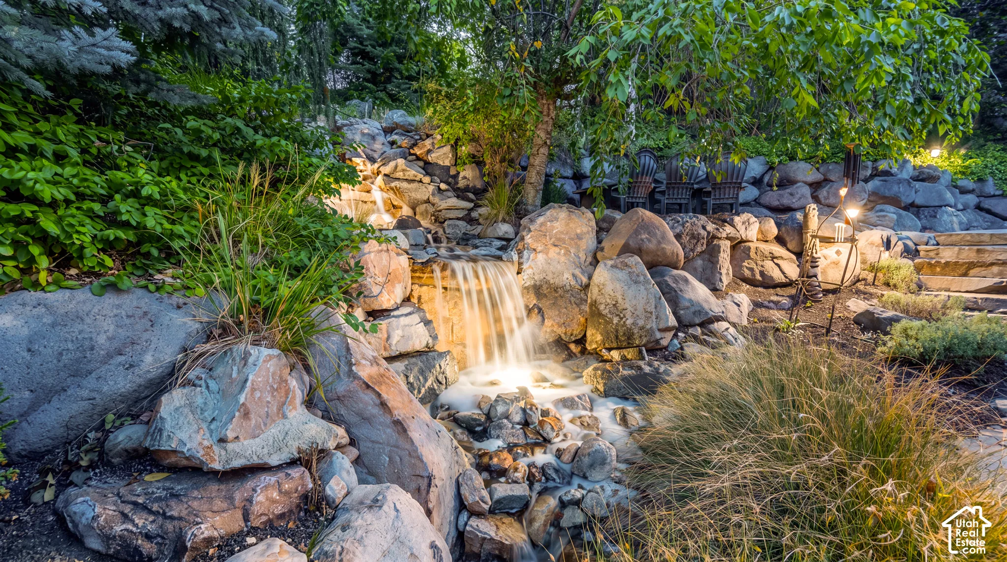 The cascading rock waterfall has integrated lighting, surrounded by beautiful landscaping and natural stone hardscaping