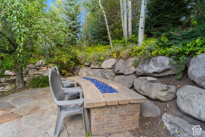 This stone patio has a built-in fire pit with blue glass, surrounded by natural rock retaining walls and mature trees, it sits next to the waterfall and overlooks the outdoor kitchen and seating areas.