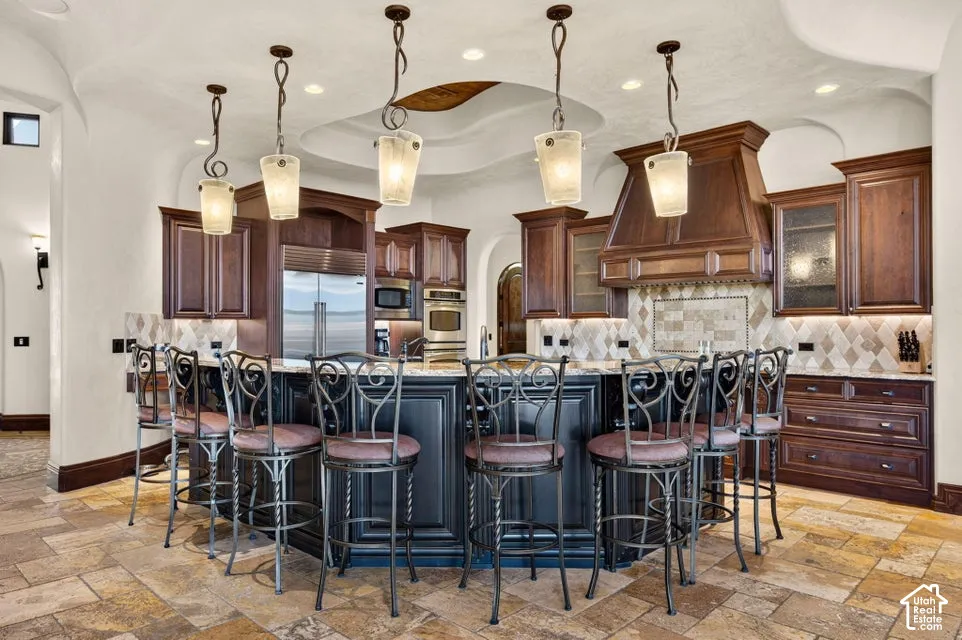 Expansive kitchen island with seating, dark wood-finish cabinetry, built-in stainless steel appliances, decorative tile backsplash, and overhead pendant lighting
