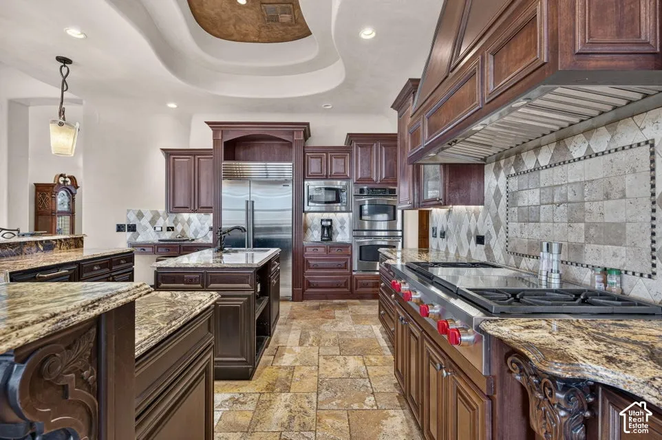 Gourmet kitchen with rich wood cabinetry, stone countertops, stainless steel appliances, a tiled backsplash, and recessed lighting