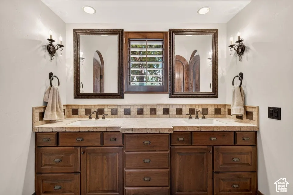 Dual sinks with bronze-finish fixtures, wood-finish cabinetry, and a tile backsplash