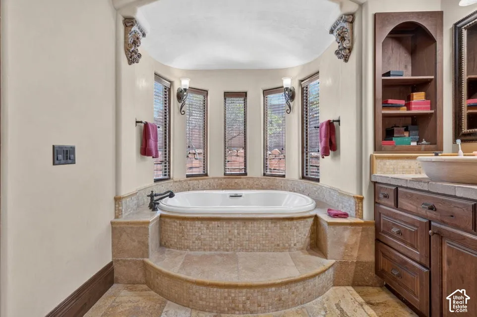 Soaking tub with mosaic tile surround, multiple windows with blinds, built-in vanity with vessel sink, and an arched built-in shelving unit