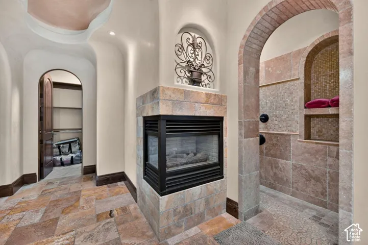 Corner fireplace with stone surround, arched doorways, tiled flooring, and a tiled shower with integrated shelving