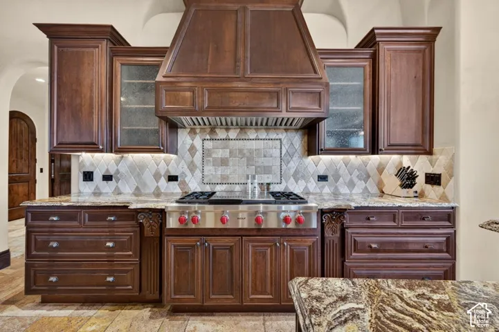 Dark wood cabinetry, built-in range hood, stainless steel gas range, patterned tile backsplash, and stone countertops