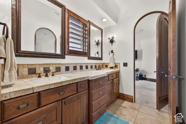 Dual vanity with individual sinks, wood-finish cabinetry, tile countertop, decorative tile backsplash, and arched entryway
