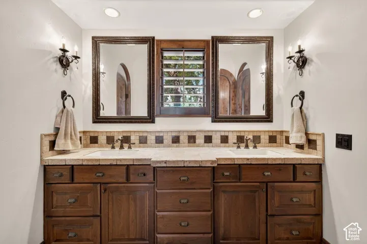 Dual sinks with bronze-finish fixtures, wood-finish cabinetry, and a tile backsplash