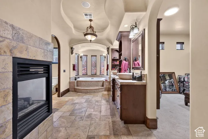 Built-in gas fireplace with stone surround, tile flooring, built-in vanity with vessel sink, built-in shelving, and a large soaking tub