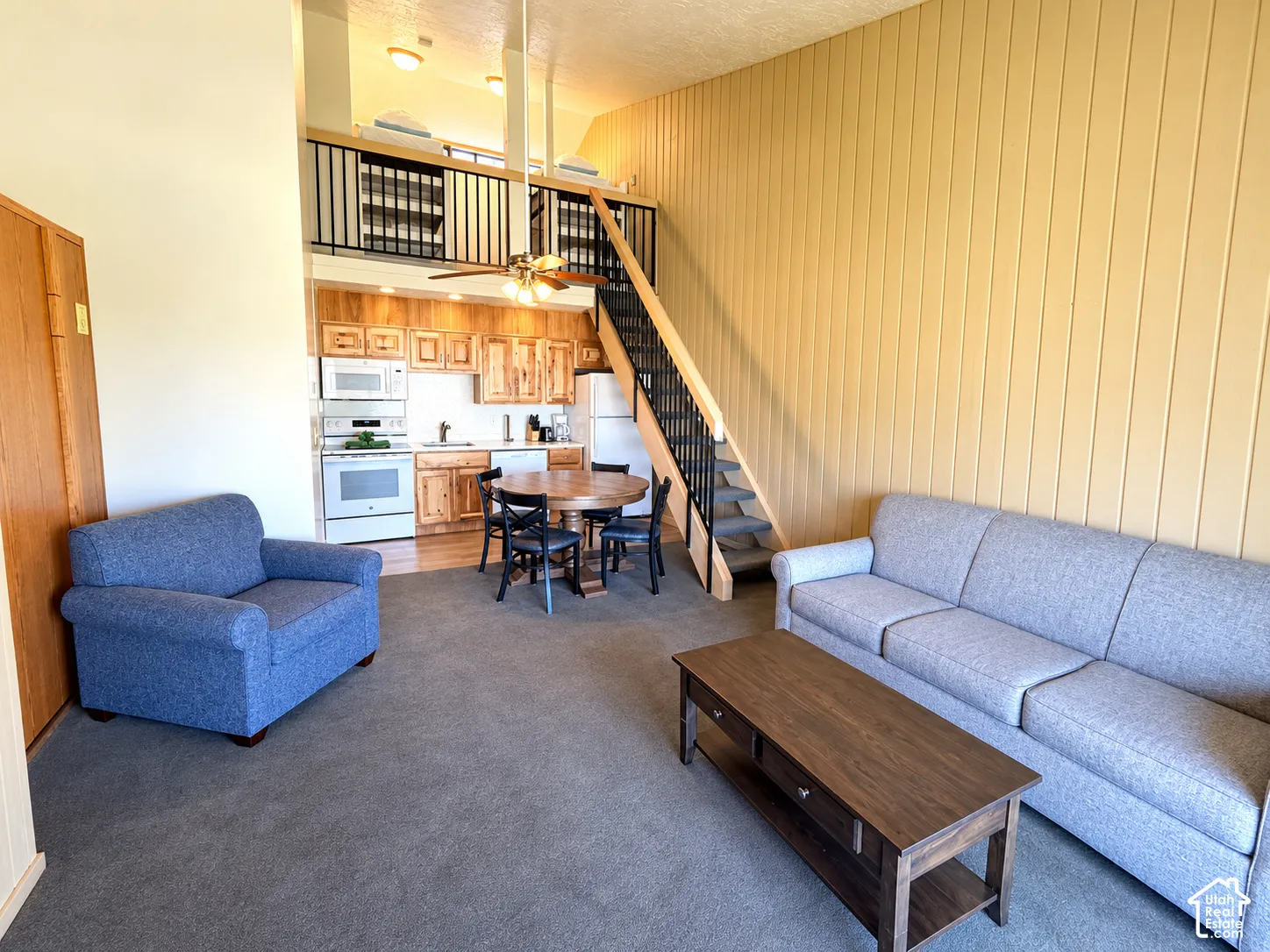 Open-concept living area featuring a kitchen with wood cabinetry and white appliances, a carpeted living space, a dining area with wood-finish flooring, and a staircase leading to an upper loft with a black metal railing