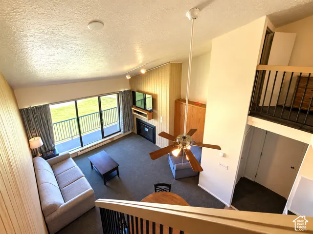 Vaulted ceiling with ceiling fan, wood-paneled accent wall, built-in fireplace with mantel, sliding glass doors to a balcony, and carpeted flooring