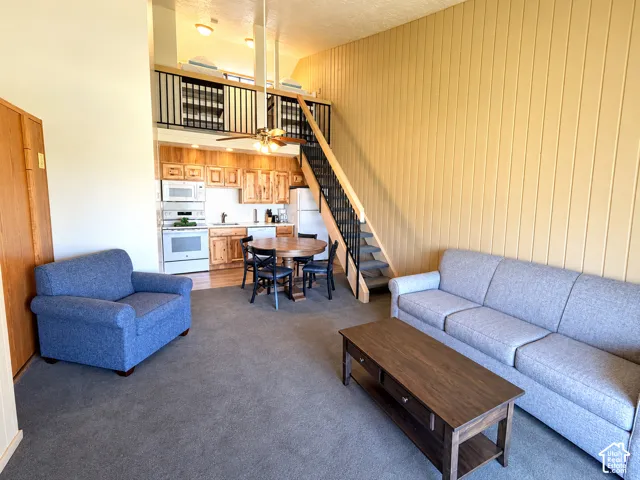 Open-concept living area featuring a kitchen with wood cabinetry and white appliances, a carpeted living space, a dining area with wood-finish flooring, and a staircase leading to an upper loft with a black metal railing