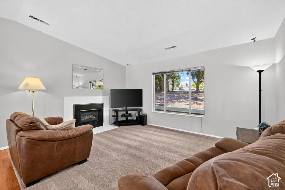 Carpeted living space with a fireplace featuring a white surround, a large window, and vaulted ceilings