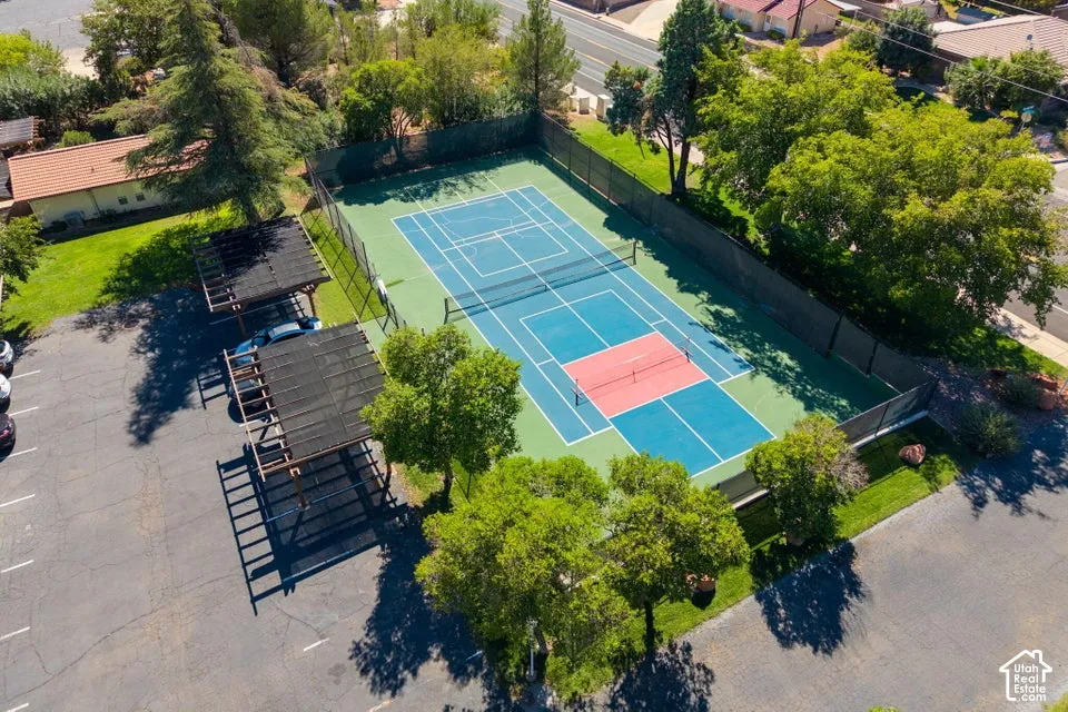 Asphalt parking area with multiple covered parking structures, a fenced tennis court, and surrounding mature trees