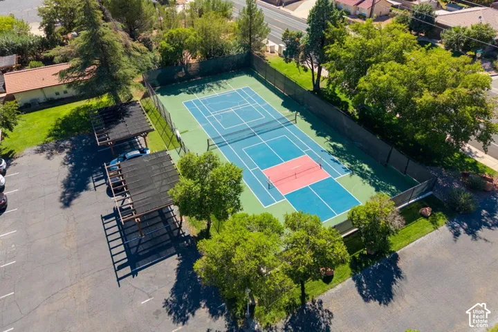 Asphalt parking area with multiple covered parking structures, a fenced tennis court, and surrounding mature trees