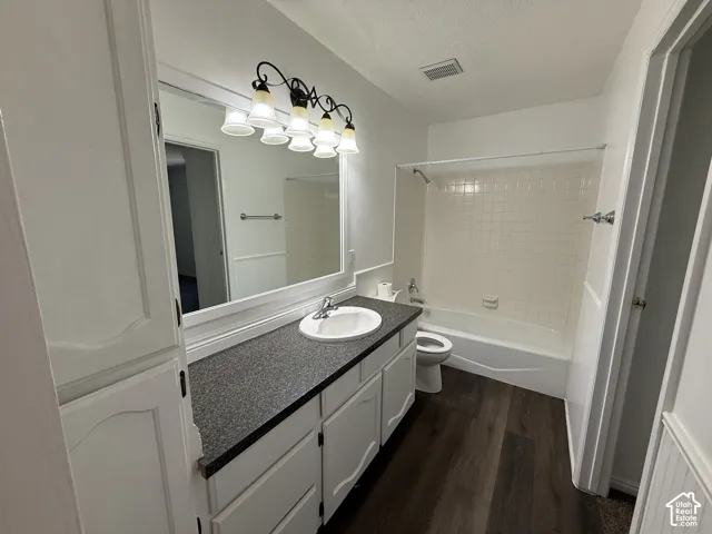 Bathroom featuring wood-finish flooring, a white vanity with an integrated sink, a dark speckled countertop, and a built-in tub with a tiled surround