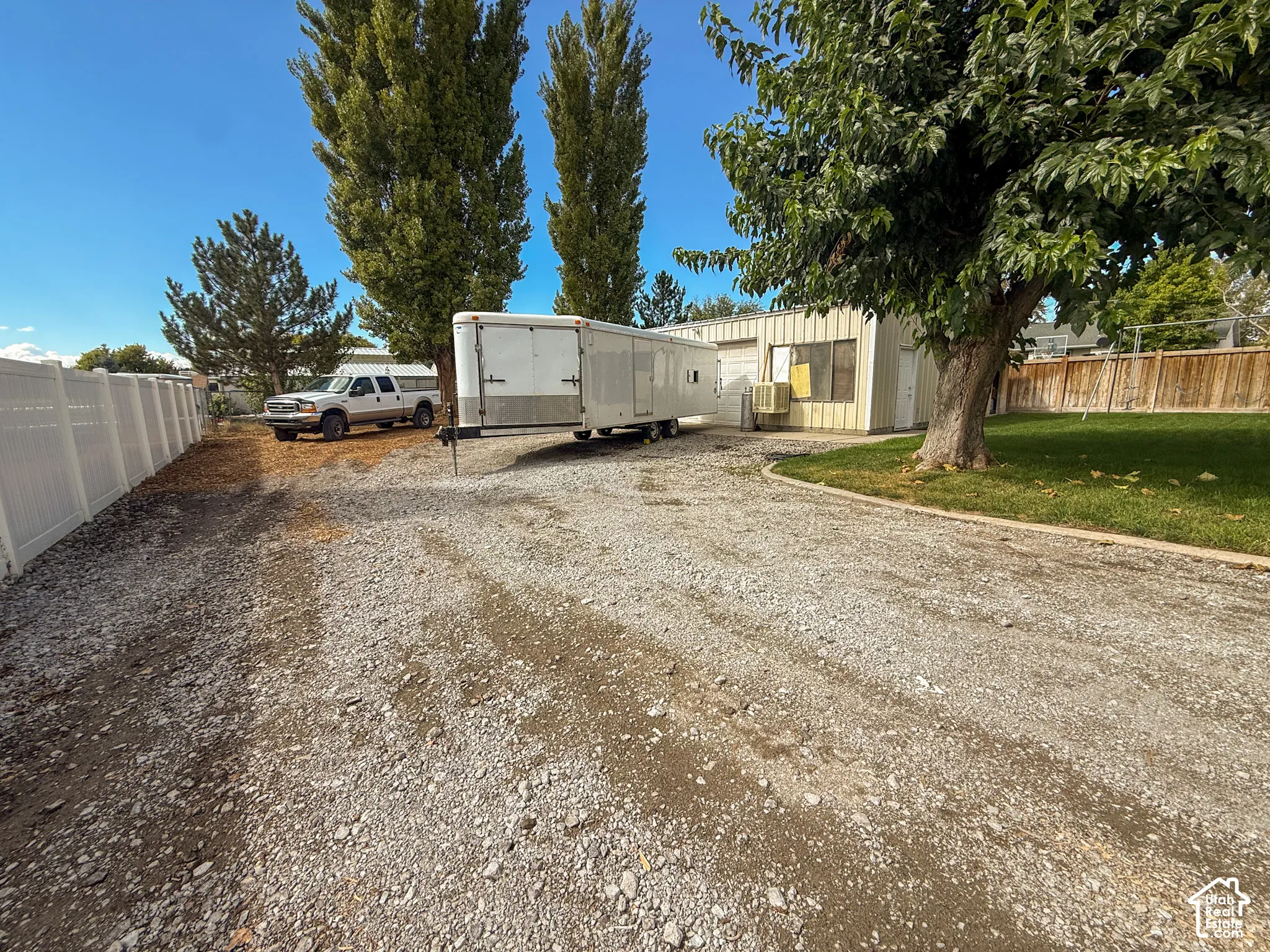 Gravel driveway providing substantial parking, white vinyl fencing, mature trees, and a wooden fence