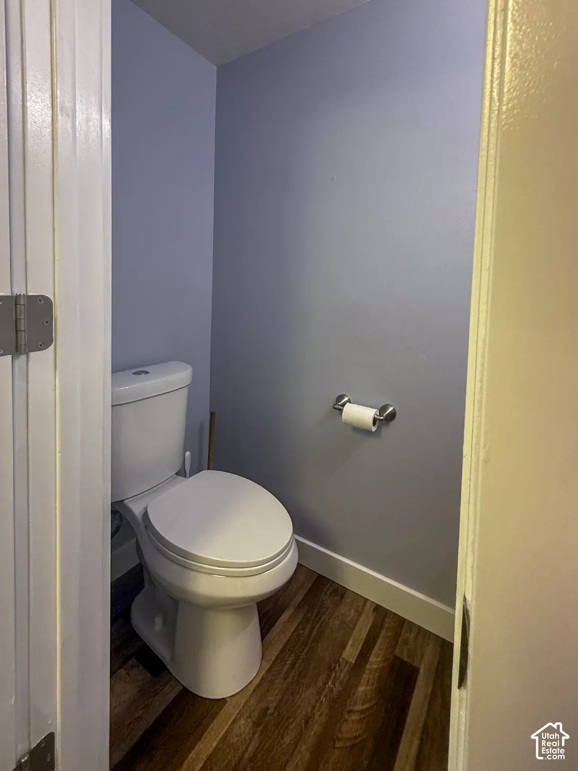 Wood-finish flooring with white baseboards, a white toilet, light blue wall paint, and a chrome toilet paper holder