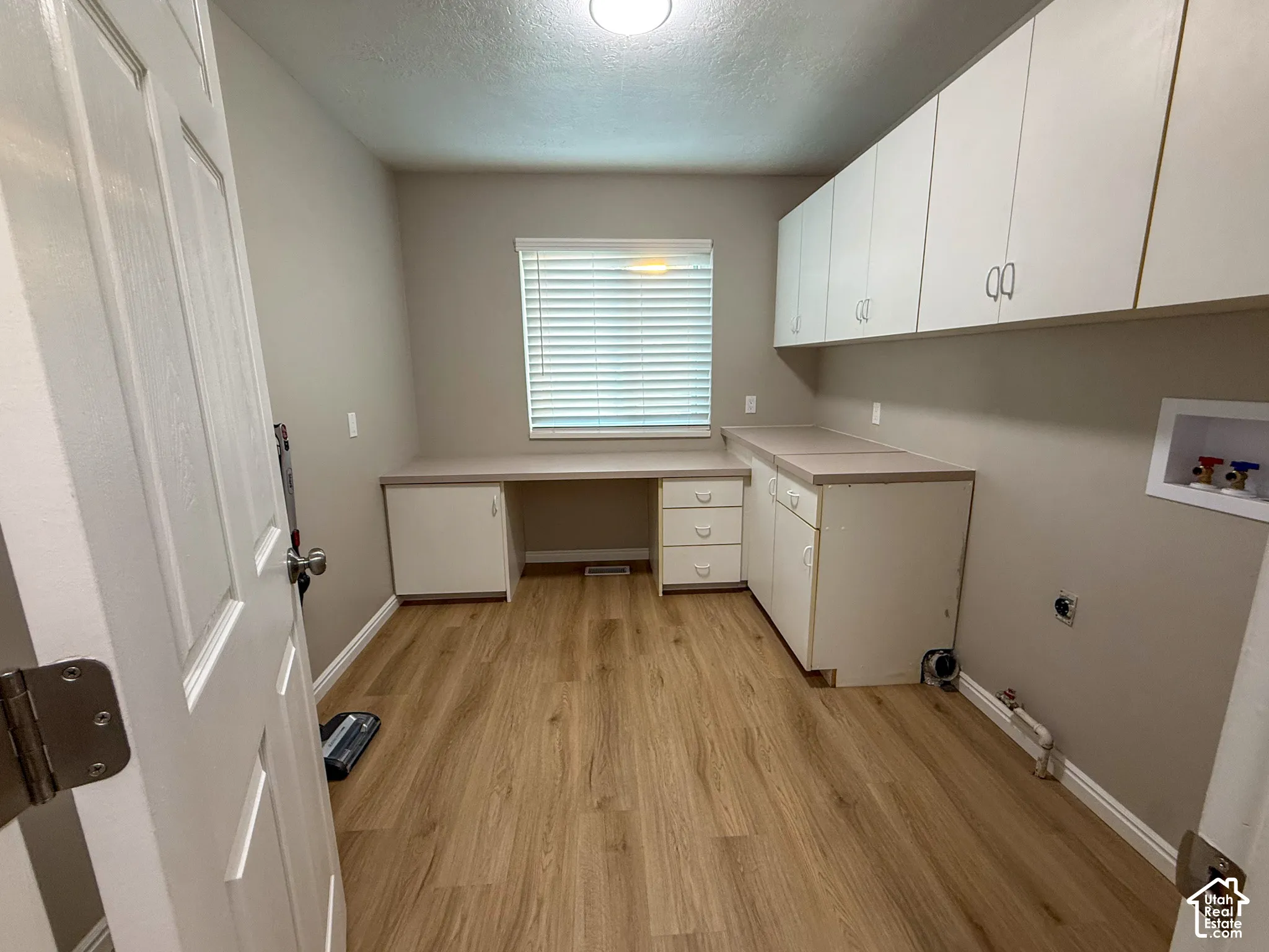 Wood-finish flooring, built-in white cabinetry, integrated desk space, upper wall-mounted white cabinets, and a window with blinds